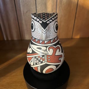 May include: A ceramic owl figurine featuring a white base with intricate hand-painted geometric patterns in black and terracotta red. The owl has large circular eyes, a pointed beak, and detailed square and triangular motifs covering its body.