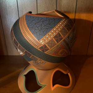 May include: A handcrafted, ceramic vessel with intricate geometric patterns in blue, brown, and red. The vessel sits atop a matching base with cut-out shapes. The design features triangles, lines, and other shapes, creating a unique decorative piece.