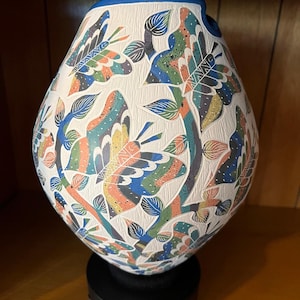 Op de afbeelding: Een druppelvormige keramische vaas met een getextureerd wit oppervlak voorzien van uitgesneden, kleurrijke vlinder- en bladmotieven in blauw, groen, oranje en zwart. De vaas heeft een effen blauwe rand en rust op een ronde zwarte voet.