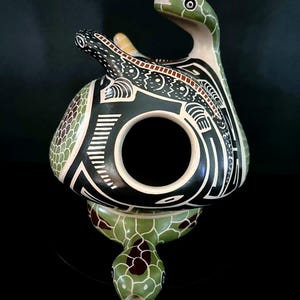 May include: A ceramic turtle sculpture with a snake-like head and neck. The sculpture is painted in green, black, and white, with detailed geometric patterns. The turtle's shell has a textured, scale-like design.