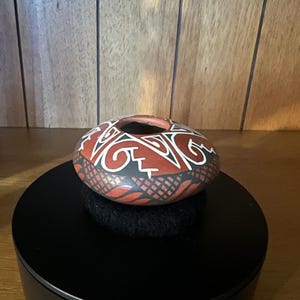 Op de afbeelding: Een kleine, lage terracotta kom met een ronde vorm en een brede opening. De buitenkant heeft een matte afwerking met ingewikkelde, met de hand geschilderde geometrische patronen in zwart en wit, waaronder kruisarceringen en golvende lijnen.
