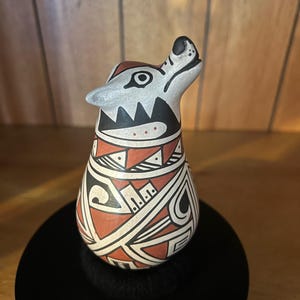 May include: A hand-painted ceramic figurine shaped like a howling wolf. The surface features a matte finish with a geometric pattern in terracotta, black, and white. The wolf has a pointed snout, alert ears, and a stylized design covering its body.