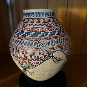 Op de afbeelding: Een ronde keramische vaas met een witte basis en handgeschilderde patronen in rood, blauw en zwart. Het ontwerp bevat horizontale banden met geometrische vormen, zigzaglijnen en spiralen, geaccentueerd door twee grote zwarte libelle-illustraties.