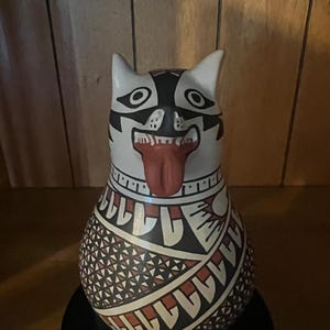 May include: A ceramic vessel shaped like a stylized animal head with pointed ears, a wide open mouth, and a protruding red tongue. The surface is decorated with intricate black, white, and terracotta geometric patterns and cross-hatch designs.