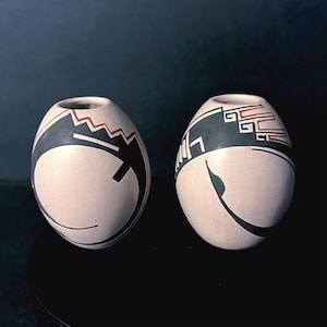 Puede incluir: Una taza de cerámica blanca con asa negra y el texto "But First, Coffee" en negrita negra. La taza se encuentra sobre un fondo borroso, sugiriendo un ambiente acogedor.