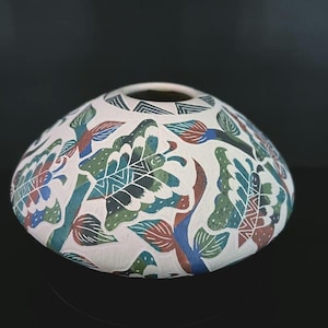 May include: A white ceramic vase with a wide, rounded form. The vase is decorated with a repeating pattern of stylized leaves and birds in shades of green, blue, and red. The design is hand-painted.
