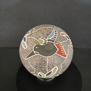 Puede incluir: Una esfera de cerámica gris moteada con un diseño de pájaro negro. El pájaro tiene detalles en rojo, azul y verde. Patrones florales blancos rodean el pájaro. La esfera se exhibe sobre un soporte negro.