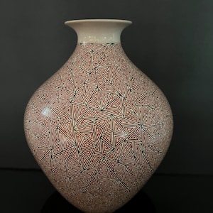 May include: A decorative ceramic vase with a cream-colored rim and a bulbous body. The vase is adorned with a repeating pattern of red and black triangles. The vase is displayed on a black surface.
