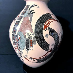May include: A white ceramic mug with a black handle and the text "But First, Coffee" in a bold, black font. The mug is set against a blurred background. The mug appears to be approximately 10 cm tall.
