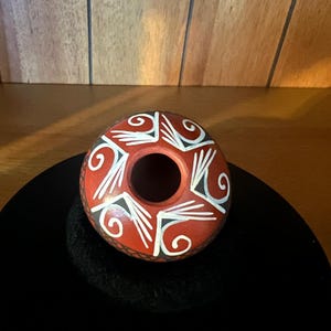 Op de afbeelding: Een kleine, ronde terracotta-kleurige kleipot met een ronde opening aan de bovenkant. De buitenkant is versierd met een herhalend patroon van witte en zwarte geometrische vormen en krullende lijnen in een radiaal ontwerp rond het midden.