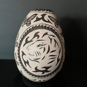 Op de afbeelding: Een eivormig, crèmekleurig decoratief object met ingewikkelde, donkerbruine gesneden ontwerpen. De patronen omvatten gestileerde vormen en een centraal cirkelvormig ontwerp. Het object wordt op een zwart oppervlak weergegeven.