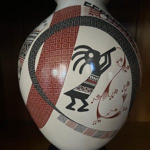 Pode incluir: Um vaso de cerâmica arredondado com base branca e padrões geométricos em preto e vermelho. O design inclui uma silhueta preta central de uma figura tocando flauta com cabelos longos, rodeada por motivos de grade e pequenas notas musicais.