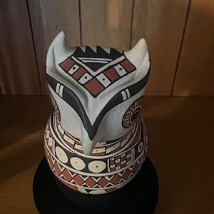 May include: A ceramic owl figurine featuring a cream-colored base with intricate geometric patterns in black and terracotta red. The owl has pointed ears, large circular eyes, and a sharp beak, with detailed line work covering its head and body.