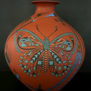 Pode incluir: Um vaso de cerâmica cor de terracota com um grande desenho de borboleta. A borboleta é delineada em preto e verde-azulado, com padrões geométricos intrincados. O vaso tem um corpo arredondado e um pescoço estreito, mostrando cerâmica nativa americana.