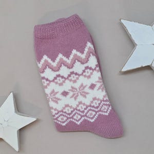 Op de afbeelding: Een gebreide sok in tinten mauve, wit en lichtroze. De sok heeft een patroon met zigzags, ruiten en sneeuwvlokachtige vormen. De boord en de teen zijn effen mauve.
