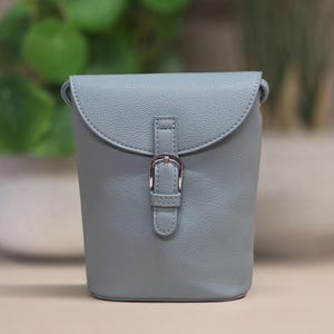 Puede incluir: Un bolso bandolera azul claro con solapa curva y cierre de hebilla plateada. El bolso tiene una superficie texturizada y un diseño sencillo y elegante. Es un bolso pequeño tipo cubo.