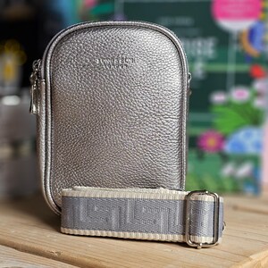 Peut inclure: Un sac à bandoulière argenté avec un extérieur en cuir texturé et une bandoulière à motif blanc et gris. Le sac a une fermeture éclair argentée et le nom de la marque "LONGCHAMP" est embossé sur le devant.