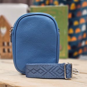Puede incluir: Un pequeño bolso bandolera de cuero azul claro con cierre de cremallera y una correa estampada. El bolso está sobre una superficie de madera.