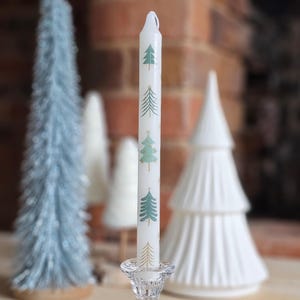 Puede incluir: Vela blanca con un diseño de árboles de Navidad verde y dorado. La vela está en un portavelas de vidrio transparente.