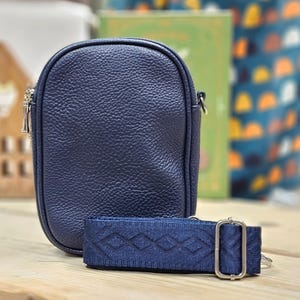 May include: A dark blue crossbody bag with a textured leather-like finish and a matching blue adjustable strap. The strap has a geometric pattern.