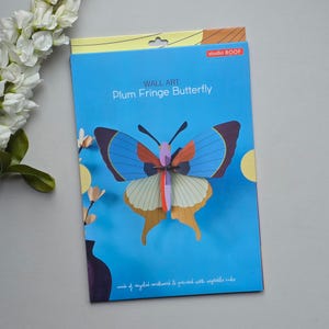 May include: A blue package featuring a colorful Plum Fringe Butterfly wall art kit. The butterfly has blue, orange, and tan wings. The package reads "WALL ART Plum Fringe Butterfly" and "studio ROOF". The product is made of recycled cardboard and printed with vegetable inks.