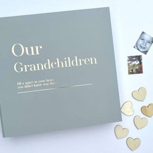 May include: A pale grey photo album with gold lettering reading "Our Grandchildren". The album features a quote and is displayed with small photographs and wooden heart decorations.