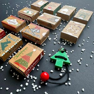 Könnte beinhalten: Eine Sammlung von Weihnachtsgeschenkboxen mit verschiedenen Designs und Texten wie "The Christmas Tree" und "Silver Bells". Ein grüner Weihnachtsbaumschmuck aus Holz mit roter Kugel und schwarzem Faden ist ebenfalls vorhanden.