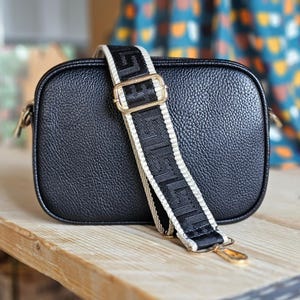 May include: Black camera bag with a white and black patterned strap. The strap has a gold buckle.
