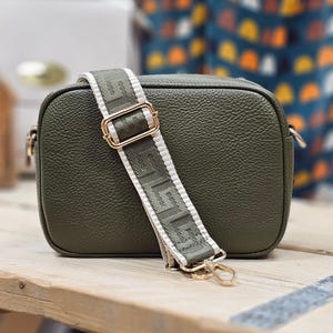 May include: Olive green crossbody bag with a textured finish. The bag features a wide, patterned strap with gold-tone hardware. The strap has a repeating geometric design in olive green and white. The bag is rectangular and has a zipper closure.