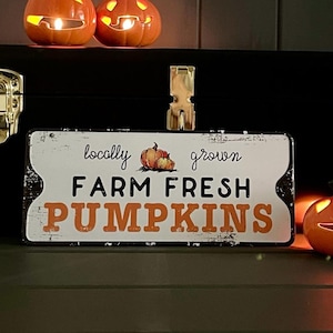 Könnte beinhalten: Ein rechteckiges Metallschild mit den Worten "locally grown FARM FRESH PUMPKINS" in Schwarz und Orange. Das Schild hat einen weißen, abgenutzten Hintergrund und einen goldenen Clip. Zwei geschnitzte Kürbisse mit brennenden Kerzen befinden sich darüber.