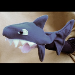 May include: A blue plush shark puppet with a white eye and a pink mouth. The puppet has a large fin on the back and a smaller fin on the side.