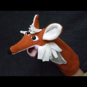 May include: A brown and white fox hand puppet with large black eyes. The puppet has a wide open mouth with a pink tongue.