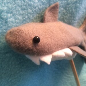 May include: A gray felt shark puppet with a white felt mouth and black bead eyes. The shark is attached to a wooden stick.