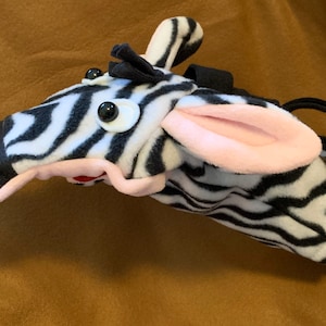 May include: A black and white zebra puppet with pink ears and large googly eyes. The puppet is made of soft, plush fabric and has a black strap for the hand.
