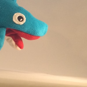 May include: A blue plush shark puppet with a red mouth and a white eye. The puppet is open-mouthed and has a white tooth visible.