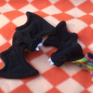 May include: Two plush black bats with rainbow fabric on the underside of the wings. One bat has white teeth and the other has white teeth and black eyes.
