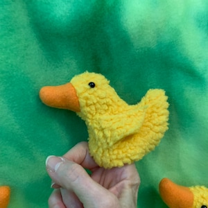 May include: A yellow fuzzy duck finger puppet with an orange beak and black eyes. The puppet is held by a hand against a green background.