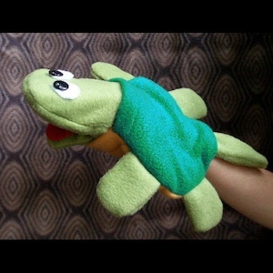 May include: Green hand puppet shaped like a turtle with a large, open mouth and googly eyes. The turtle has a green shell and yellow feet.
