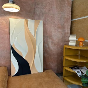 Pu&ograve; includere: Un'opera d'arte murale astratta bianca, beige e nera con un effetto 3D. L'opera &egrave; appesa a una parete con pannelli di legno color legno chiaro.