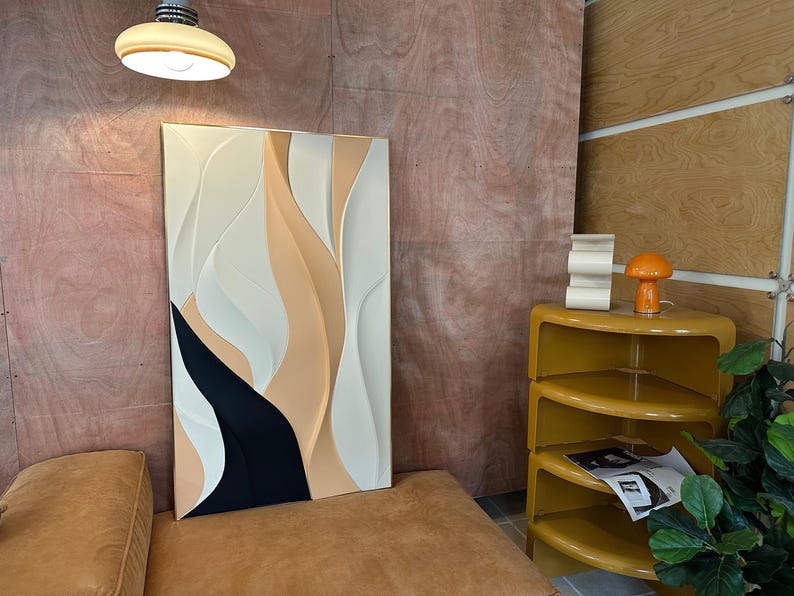 Pu&ograve; includere: Un'opera d'arte murale astratta bianca, beige e nera con un effetto 3D. L'opera &egrave; appesa a una parete con pannelli di legno color legno chiaro.