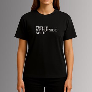 Puede incluir: Camiseta negra con el texto blanco "THIS IS MY OUTSIDE SHIRT." centrado en el pecho. La camiseta tiene mangas cortas y cuello redondo. La modelo lleva pantalones negros.