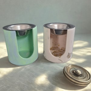 Peut inclure: Deux bougeoirs en béton avec inserts en acier inoxydable, l'un vert et bleu, l'autre rose. Une toupie en métal argenté est à côté des bougeoirs. Les bougeoirs ont une forme rectangulaire arrondie.