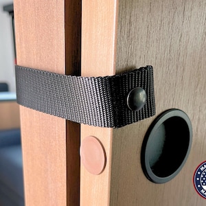 May include: A black nylon strap with a metal snap fastener secures two light brown wood-grain panels together. A circular black recessed handle is visible on the right panel, and a small circular tan bumper is attached to the left panel edge.