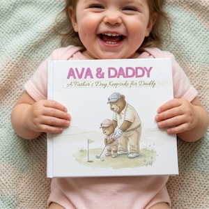May include: A child smiles while holding a book titled "AVA & DADDY" with the subtitle "A Father's Day Keepsake for Daddy." The book cover features an illustration of two bears playing golf. The background is a soft, neutral color.