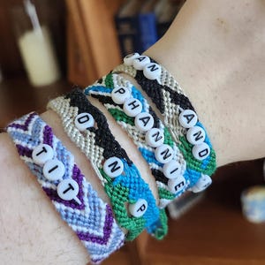 May include: Close-up of several handmade friendship bracelets. Each bracelet is a different color and pattern, with white letter beads spelling out names. The bracelets are made of woven threads in shades of blue, purple, green, black, and white.