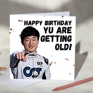 May include: A white birthday card with the text "HAPPY BIRTHDAY YU ARE GETTING OLD!" features a smiling person pointing. The person is wearing a white racing suit with black and blue accents. Confetti is scattered around the card.