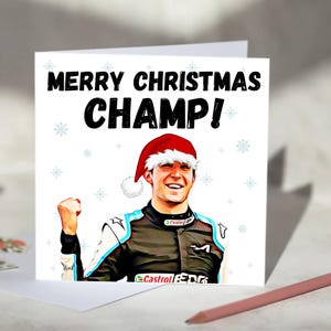 Op de afbeelding: Een witte kerstkaart met de tekst "MERRY CHRISTMAS CHAMP!" in het zwart. De kaart toont een portret van een lachende persoon met een kerstmuts en een racepak. Sneeuwvlokillustraties zijn verspreid rond de tekst.