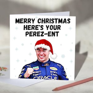 Könnte beinhalten: Weiße Weihnachtskarte mit dem Text "MERRY CHRISTMAS HERE'S YOUR PEREZ-ENT" in Schwarz. Die Karte zeigt eine Illustration eines lächelnden Mannes mit Weihnachtsmütze und blauem Rennanzug. Schneeflocken-Illustrationen sind um den Text herum verstreut.