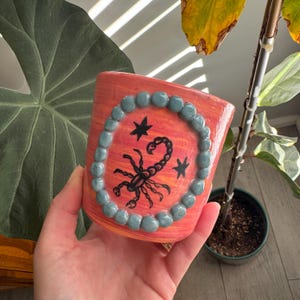 May include: A cylindrical ceramic planter with a marbled orange and pink finish. The front features a black scorpion illustration flanked by two small stars, all encircled by a raised border of smooth, round, sage green clay beads.