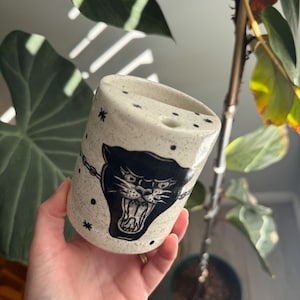 May include: A speckled off-white ceramic mug featuring a black panther illustration with an open mouth and bared teeth. The design includes a black chain graphic extending from the panther, accented by small black star and dot motifs.